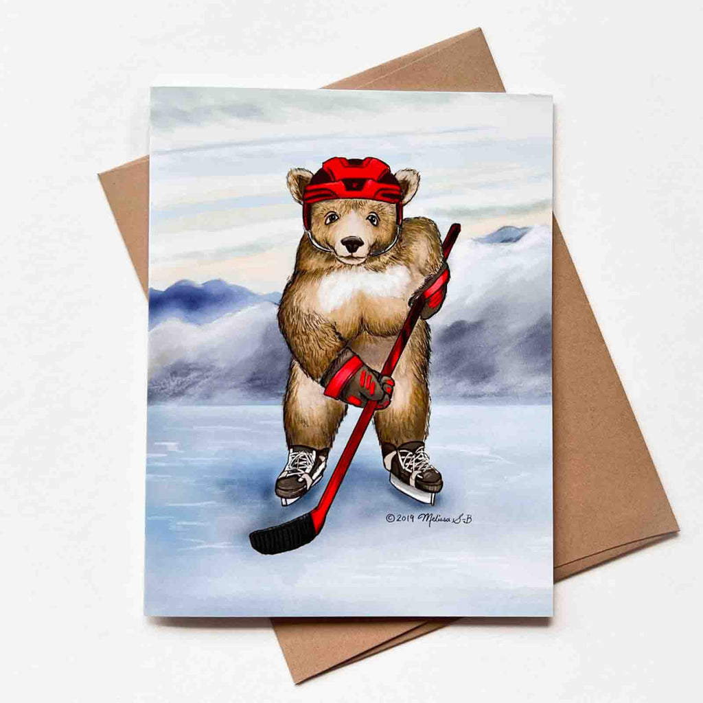 a greeting card featuring a bear playing hockey on a frozen lake against mountains in the background