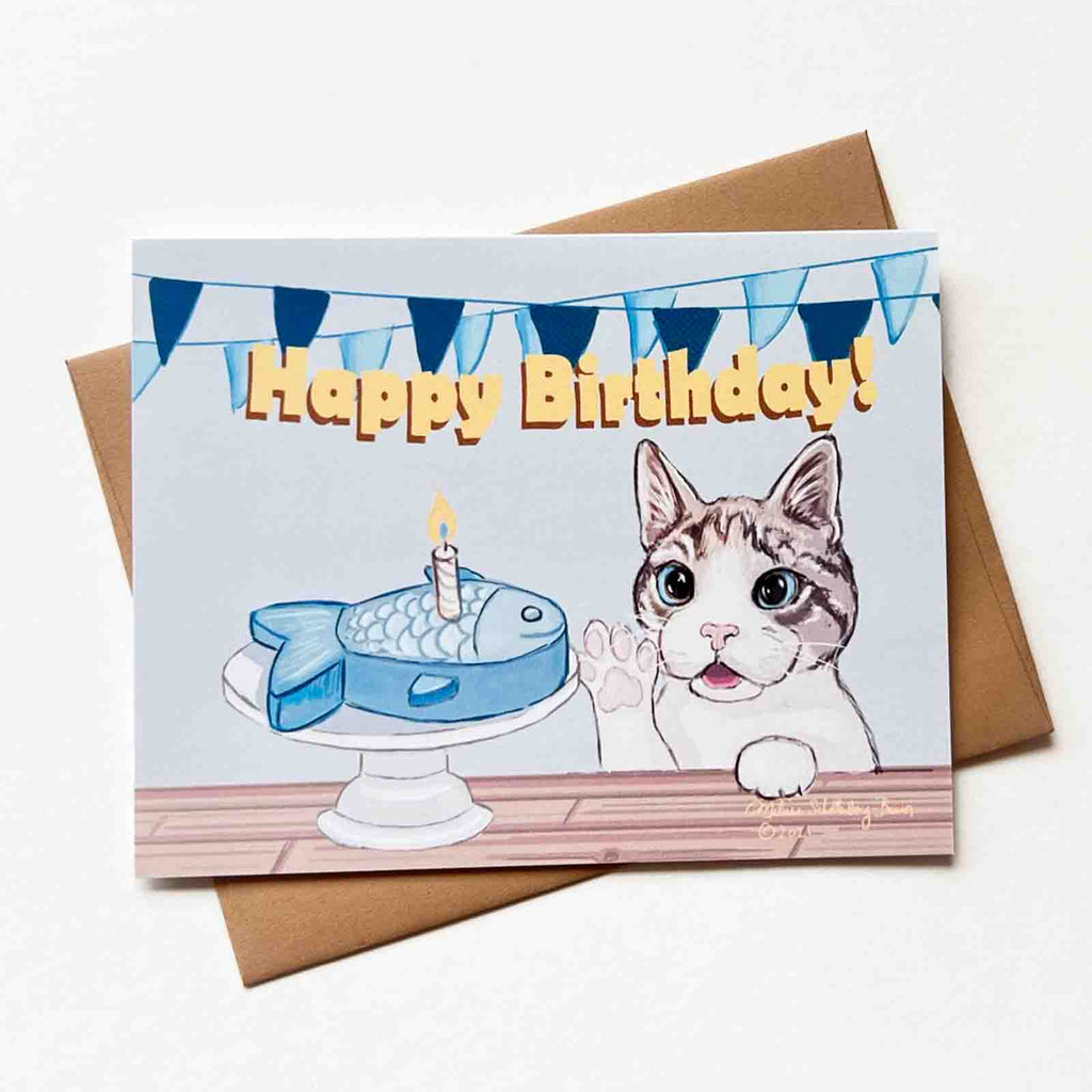 This greeting card shows a grey and white cat reaching out to his fish shaped birthday cake