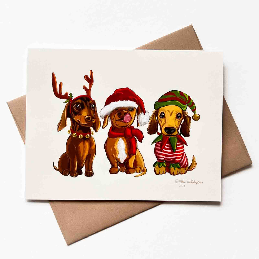 Three cute dachshund dogs wearing festive clothing is featured on this holiday card