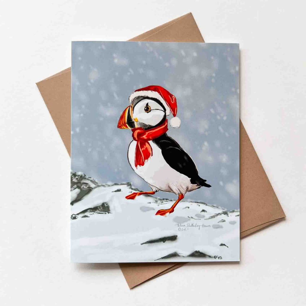 this cute holiday card features a puffin is trudging through the snow while waring a red Santa Hat.