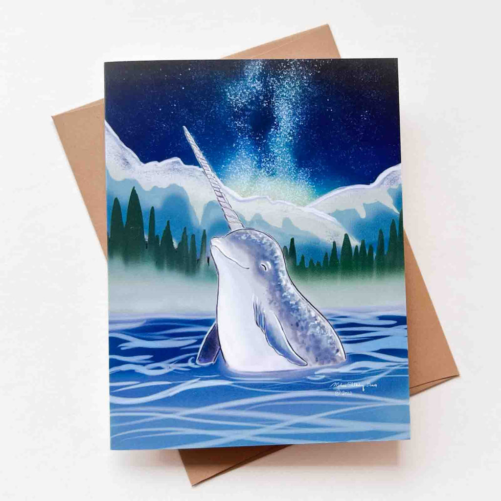 This greeting card features a narwhal coming out of the ocean at night against a backdrop of the milky way galaxy.