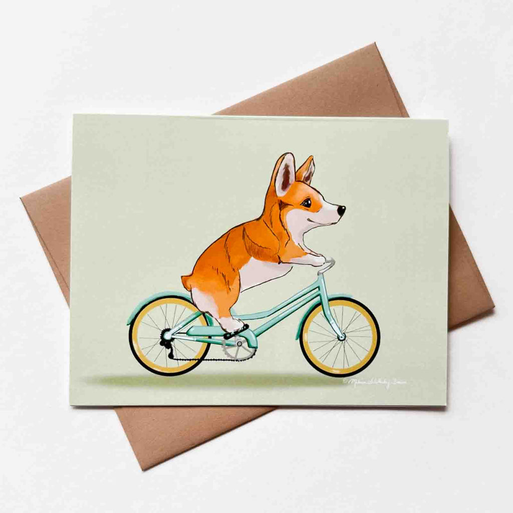 This whimsical greeting card features an orange Corgi dog riding a teal colored bike