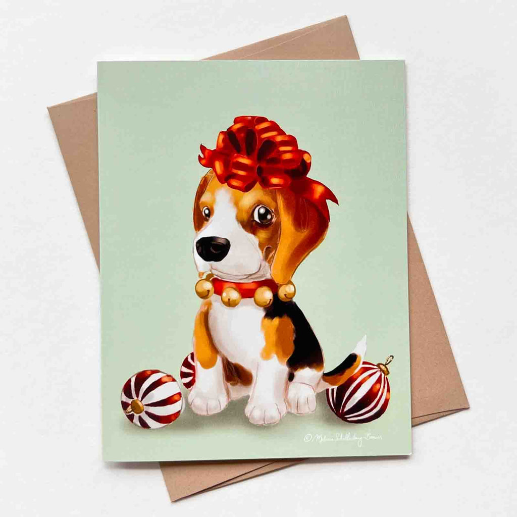 This holiday greeting card features a Beagle Puppy dog with a bow on top of its head, sitting next to Christmas ornaments and ribbons. 