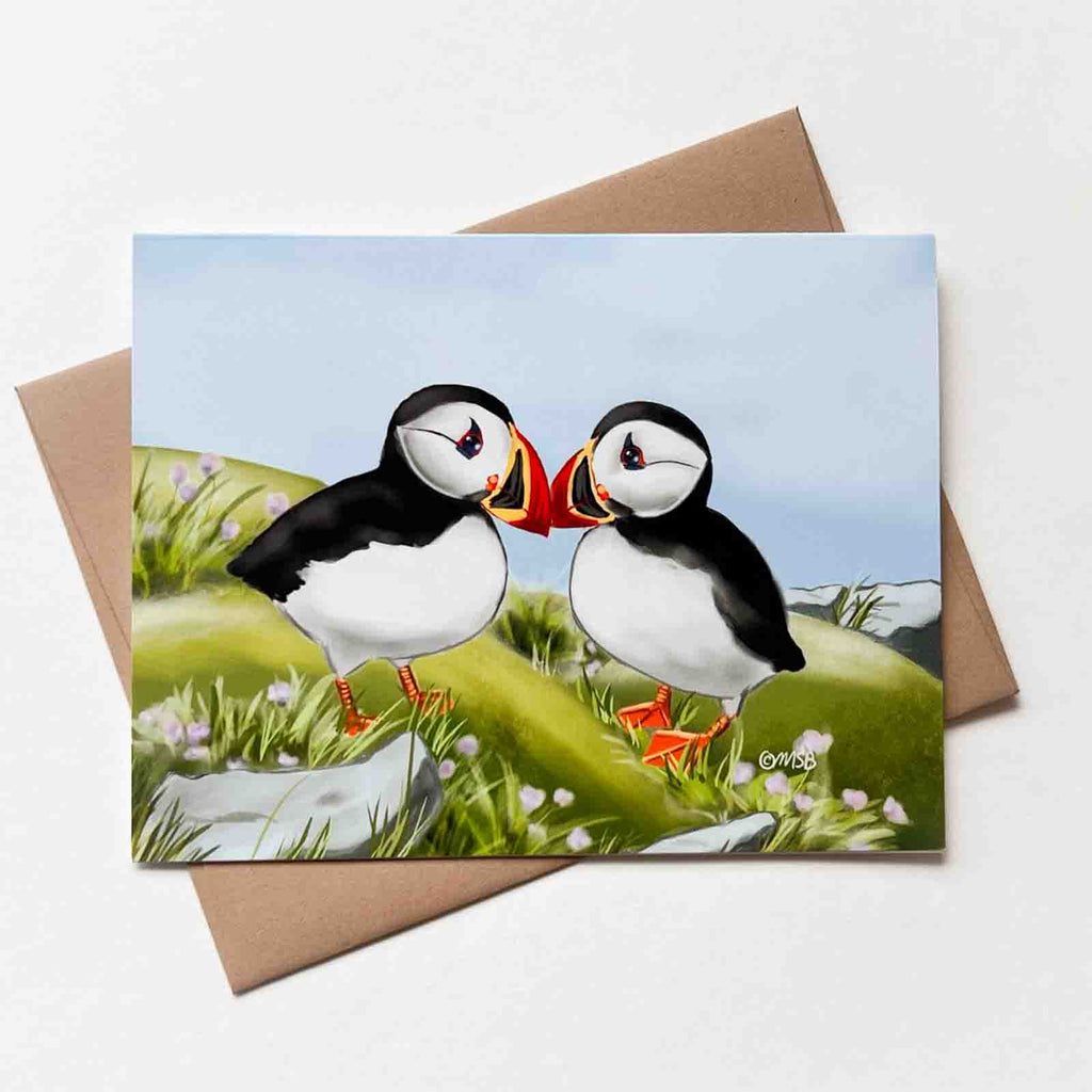 This love card features two cute little puffins facing each other while standing in a grassy field with some flowers.