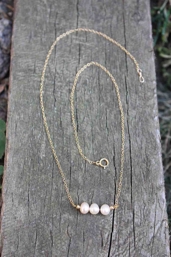 A necklace with 3 creamy coloured pearls on a dainty gold chain resting on a wooden board