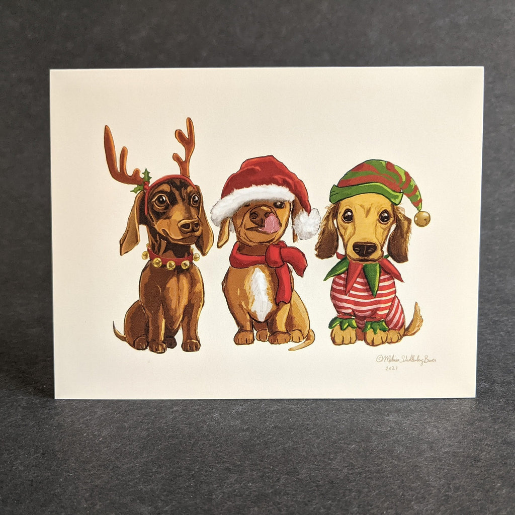 Three cute dachshund dogs wearing festive clothing is featured on this holiday card