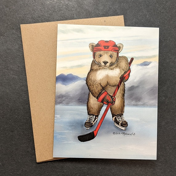 a greeting card featuring a bear playing hockey on a frozen lake against mountains in the background